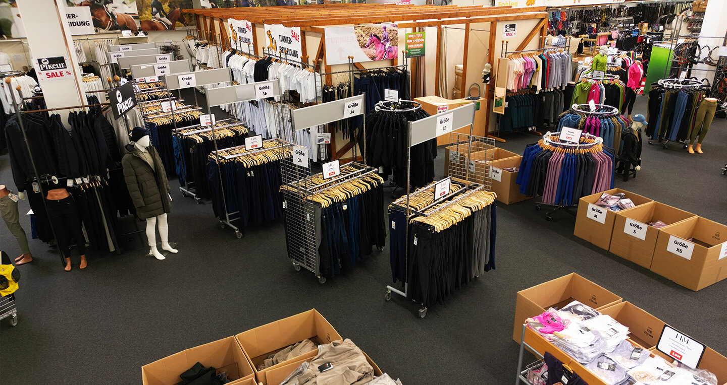 Zu sehen ist die Kleidungsabteilung. Auf dem dunklen Teppichboden stehen mehrere Rundständer mit bunten Shirts, Westen und Reithosen. Im Hintergrund sind die Umkleidekabinen zu erkennen.